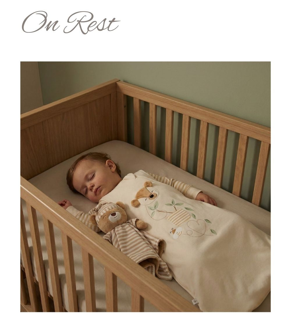 Sleeping baby in a wooden cot wearing a neutral-toned sleep sack with a soft toy beside them.