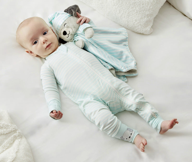 A baby lies on a white surface, wearing Zelkii's blue and white striped puppy pyjamas and holding the matching 'asleep' puppy LuviDuvi lovey.