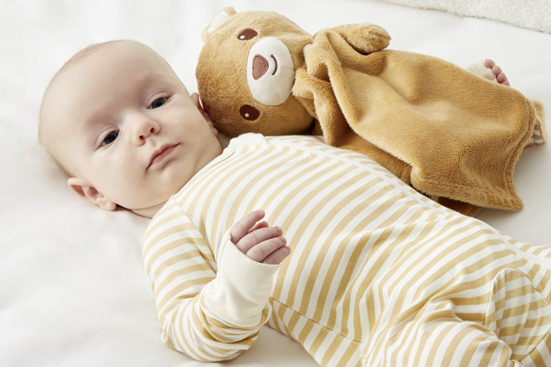 A baby lying on a white surface, next to the 'awake' Zelkii teddy bear LuviDuvi lovey.